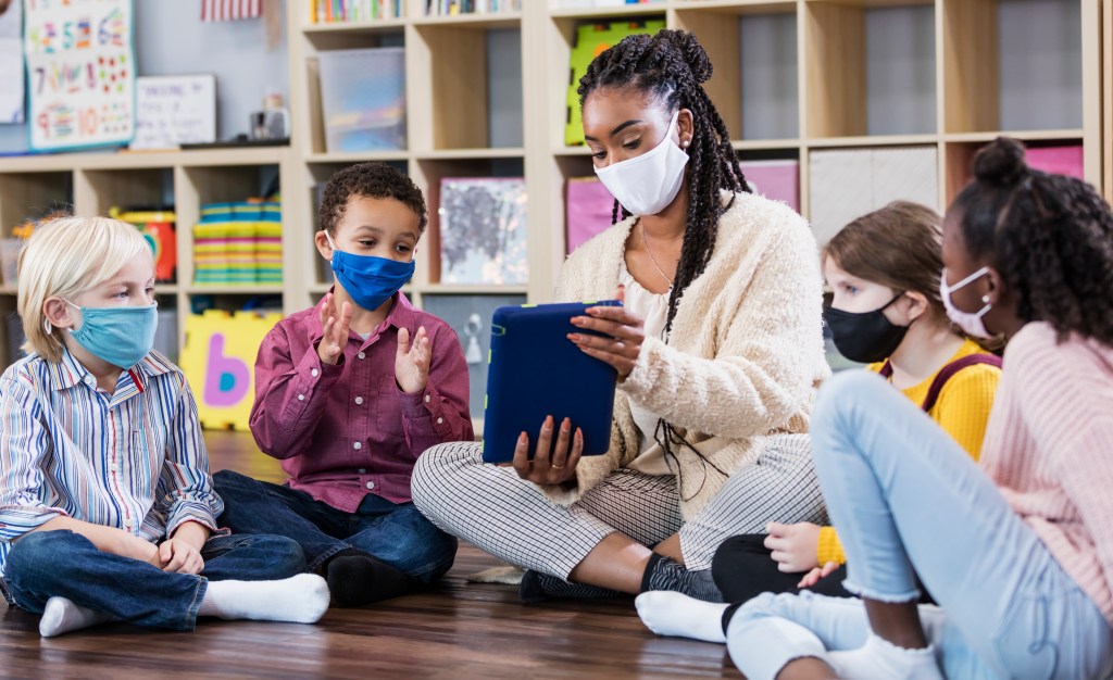 A multi-ethnic group of kindergarten or first grade students and their teacher in the classroom, sitting together in a circle on the floor. The children are 5 to 7 years old. They are all wearing masks, back to school during the COVID-19 pandemic, trying to prevent the spread of coronavirus. The teacher, an African-American woman, is holding a digital tablet and a boy is looking at it while clapping his hands.