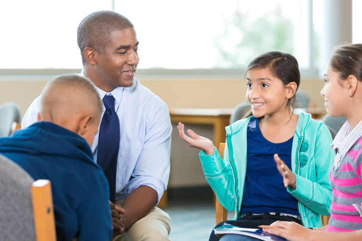 Diverse group of elementary age little girls and little boys are sitting in circle with mid adult African American male counselor or therapist. Mental health professional is leading children's group therapy session. Children are smiling and talking expressively.