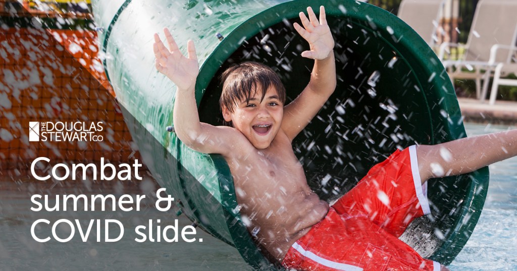 A young boy is sliding out of a water slide in red swim shorts. 