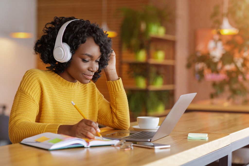 Smiling black teacher with wireless headset studying online, using laptop at cafe, taking notes