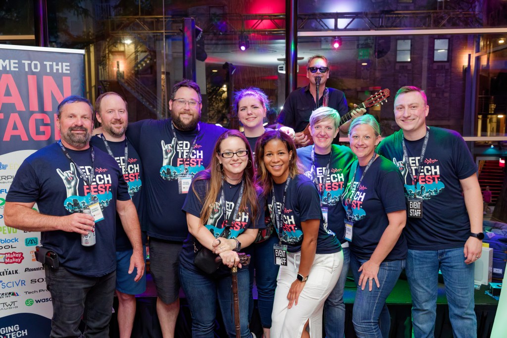 Team of colleagues in matching t-shirts posing together for a photo in front of a stage at industry event